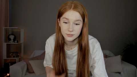 Media: Video of a young, pale-skinned, red-haired woman with long, straight hair in a white t-shirt, sitting on a bed with pink pillows and a white sheet in a dimly lit room.