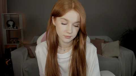 Media: Video of a fair-skinned, long-haired redhead woman with closed eyes, wearing a white shirt, seated in a dimly lit living room with a gray couch, white curtains, and a lamp.