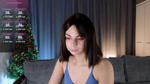 Media: Video of a young woman with straight, shoulder-length dark hair, wearing a blue tank top, seated on a grey couch. Christmas tree with lights and red ornaments in the background.