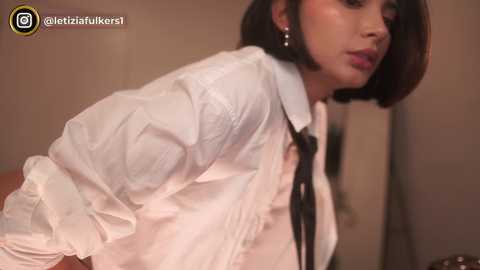 Media: A video of a woman with medium skin tone, bobbed black hair, and wearing a white button-up shirt with a black tie, standing indoors with a soft-focus background.