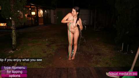 Media: Video of a nude woman with long dark hair standing in a muddy yard, holding a green towel.