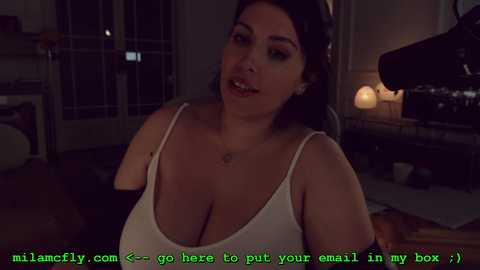 Media: A video shows a curvy woman with dark hair, wearing a white tank top, sitting in a dimly lit living room. Text reads, \"mila.mcfly.com <3 go here to put your email in my box,\" suggesting a viral meme.