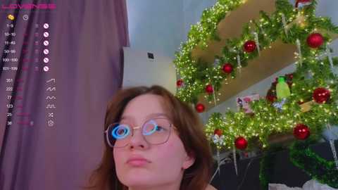 Media: Video of a young woman with glasses and brown hair, standing in a room decorated with a Christmas wreath and red ornaments.