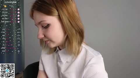 Media: A video of a young woman with light skin and shoulder-length blonde hair, wearing a white blouse, looking down at a phone displaying a \"Loen\" app screen with cryptocurrency data.