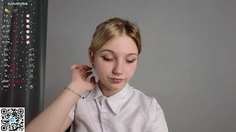 Media: Video of a young Caucasian woman with blonde hair in a white shirt, adjusting her hair in front of a gray wall with a digital screen displaying a menu.