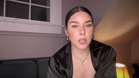 Media: Video of a fair-skinned woman with straight black hair, wearing a black satin robe and gold necklace, sitting indoors next to a window with a white frame, and a yellow lamp on the right.