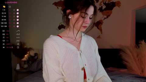 Media: Video of a fair-skinned woman with dark hair, wearing a white ribbed cardigan and red top, in a dimly lit bedroom with a world map and potted plants in the background.