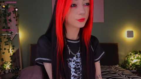 Media: Video of a pale-skinned person with long, vibrant red hair and black clothing, including a graphic t-shirt, in a dimly lit room with green walls, a bed, and a zebra-print pillow.