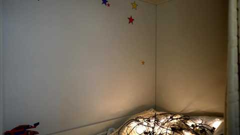 Media: A video of a cozy corner of a room with a white wall adorned with colorful star stickers and a small, dimly lit window. A bed with a cream-colored blanket and intricate metal bedframe is partially visible.