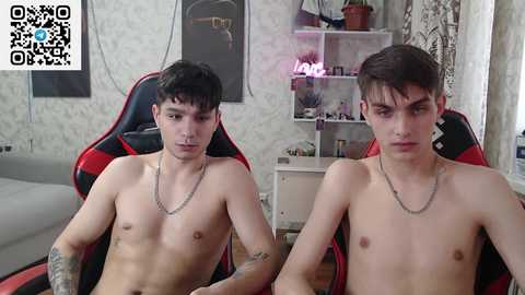 Media: Video of two young, shirtless, slender men with pale skin and dark hair, seated on red and black gaming chairs in a dimly lit, cluttered room with white walls and shelves.