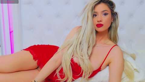 Media: Video of a young, attractive woman with long, blonde hair, fair skin, and red lipstick, lying on a white bed in a modern, minimalist room. She wears a revealing red dress.