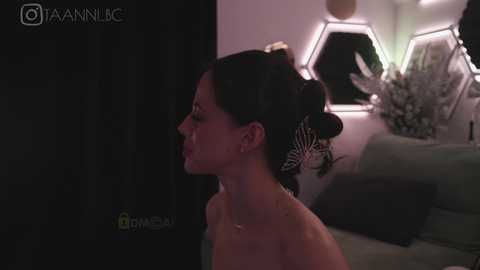 Media: A video of a woman with dark hair styled in a bun, wearing a butterfly hairclip, standing nude in a dimly lit room with modern decor.