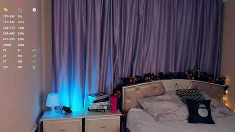 Media: Video of a dimly lit bedroom with a purple curtain backdrop, a beige bed with white sheets and pillows, and a white nightstand with books and a lamp emitting a blue glow.