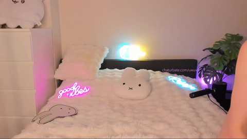 Media: A cozy, pink-lit bedroom with a white bed, a plush white bunny, and a neon \"good vibes\" sign. A green potted plant and a Star Wars helmet add a whimsical touch.