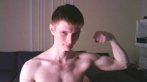 Video of a young, shirtless, muscular man with short brown hair flexing his right arm, against a dark couch and beige wall with vertical blinds.