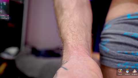 Media: Video of a close-up of a person's forearm with a tattoo, holding a smartphone, partially visible. The background is blurred, showing a dark room with some indistinct objects. The person is wearing a grey shirt with blue accents.