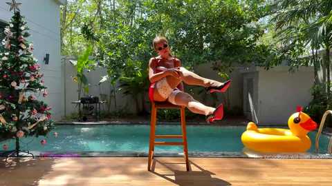 Media: Video of a woman in red lingerie and high heels, seated on a stool by a pool, wearing sunglasses and a Santa hat, with a yellow rubber duck and a decorated Christmas tree in the background.