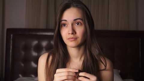 Media: Video of a young woman with long brown hair, light skin, and a neutral expression, sitting on a bed with a dark headboard, in a dimly lit room.