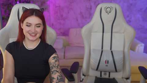 Media: Video of a smiling, tattooed young woman with red hair and cat ears, seated in a white gaming chair in a purple-lit room.