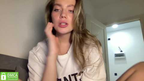 Media: Video of a young, fair-skinned blonde woman with shoulder-length hair, wearing a white t-shirt, touching her face, indoors with a white wall and door.