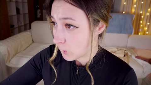 Media: Video of a pale-skinned woman with blonde hair in a bun, wearing a black zip-up top, looking shocked and mouth agape, in a cozy living room with white furniture and string lights.