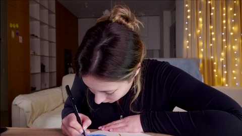 Media: Video of a young woman with light skin and brown hair in a messy bun, writing in a notebook, wearing a black long-sleeve top. Background features a cozy living room with white curtains and a soft light string.