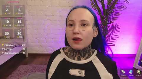 Video of a woman with pale skin, blue hair, and extensive tattoos, wearing a black bodysuit with white accents, in a modern room with white brick wall, purple lighting, and a plant.