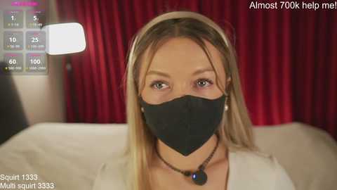 Media: Video of a blonde woman in a black face mask, white top, and necklace, against red curtains, with a digital overlay of \"Almost 100% help me\" and \"Squirt 1333\" text.