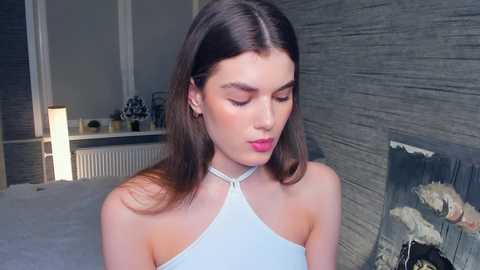 Media: Video of a young Caucasian woman with long brown hair, wearing a light blue halter top, in a modern bedroom with gray textured walls, a bed, and a lit fireplace.