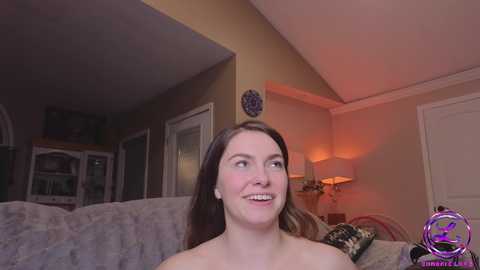 Media: Video of a smiling, topless, fair-skinned woman with long brown hair, standing indoors in a warmly lit living room with beige walls, a mirror, and a couch.