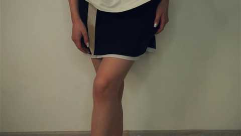 Media: Video of a person wearing a white blouse and black athletic shorts with white trim, standing against a plain, light-colored wall, emphasizing a slender leg.