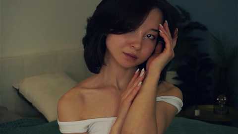 Media: Video of a young Asian woman with shoulder-length black hair, pale skin, and a slender physique. She wears a white off-shoulder top, her hands gently touching her face, smiling softly. The background shows a beige wall, a green plant, and a wooden nightstand.