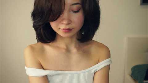 Media: A video of a young Asian woman with short, wavy brown hair, wearing a white off-shoulder knit top. She has a smooth, fair complexion and is looking down. The background is a plain, light-colored wall.