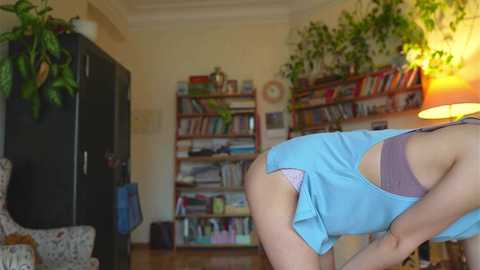 Media: Video of a woman bending over in a cozy living room, wearing a light blue tank top and revealing pink underwear. Bookshelves filled with books and potted plants surround her.