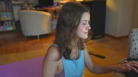 Media: Video of a young Caucasian woman with wavy brown hair in a blue tank top, sitting on a yoga mat in a cozy living room with wooden floors, bookshelves, and a couch.