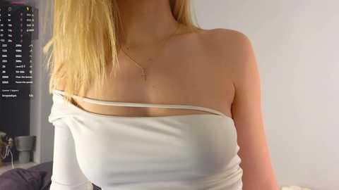 Media: Video of a young woman with fair skin and long blonde hair, wearing a white off-shoulder top that reveals her cleavage, in a dimly lit room with a black wall calendar and a desk visible in the background.