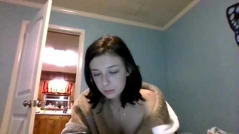 Media: A video shows a young woman with shoulder-length black hair, wearing a beige bathrobe, in a dimly lit bathroom with light blue walls and a wooden cabinet visible in the background.