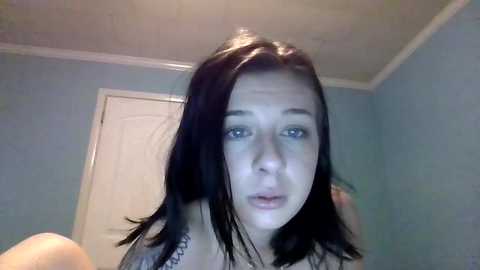 Media: A video of a young woman with straight, shoulder-length dark hair, pale skin, and blue eyes, wearing a gray top. The background features a light blue wall with a white door.