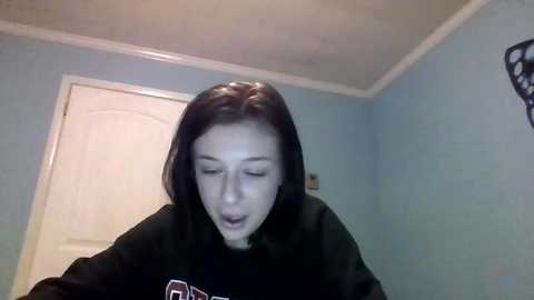 Media: A video of a young woman with straight, shoulder-length black hair, wearing a black sweatshirt, eyes closed, in a blue-painted room with a white door and clock.