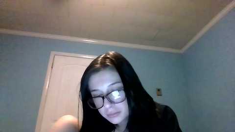 Media: Video of a young woman with long black hair, wearing glasses, in a room with light blue walls and white trim. She appears to be in a bathroom, looking down.