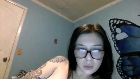 Media: A video of a young woman with straight black hair, glasses, and a tattoo on her arm, leaning forward in a room with light blue walls and a butterfly wall decal.