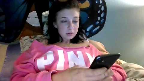 Media: Video of a young woman with fair skin and long black hair, wearing a pink sweatshirt with \"PINK\" in white letters, focused on a black smartphone. She lies on a bed with a colorful, patterned blanket.