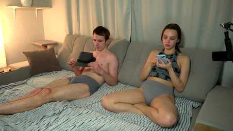 Media: Video of a light-skinned man and woman lying on a grey couch, wearing grey underwear, playing video games on a Nintendo Switch. Background features a striped blanket and a lamp.