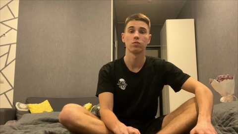 Media: Video of a young Caucasian man with short brown hair, sitting cross-legged on a bed in a modern bedroom with gray walls, a white door, and geometric-patterned wallpaper.