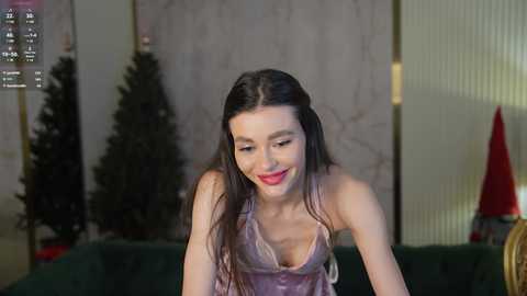 Media: Video of a fair-skinned woman with long dark hair, wearing a pink satin camisole, sitting on a green couch, surrounded by Christmas trees and a red cone-shaped object in a dimly lit room.