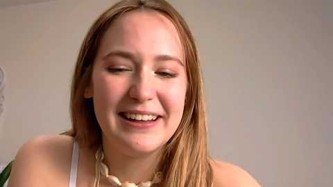 Media: Video of a young Caucasian woman with long, straight, light brown hair, wearing a white tank top and a chunky, beige necklace. She has a gentle smile, light skin, and is standing against a plain, light-colored background.