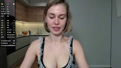 Media: A video of a fair-skinned woman with a short bob haircut, wearing a revealing black dress with white lace trim, in a modern kitchen with wooden cabinets.