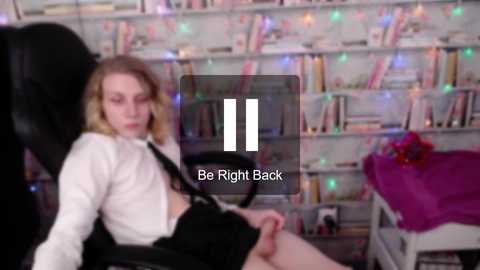 Media: Video of a young man with shoulder-length blonde hair in a white shirt, black tie, and black skirt, sitting in a black office chair, in a colorful, cluttered bookshelf-filled room. Text overlay: \"Be Right Back.\