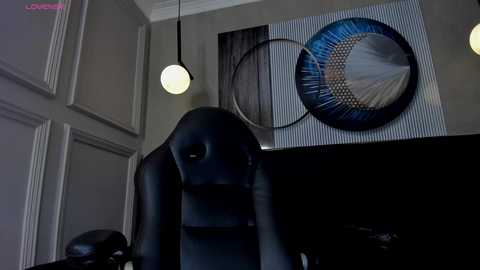 Media: Video of a black leather gaming chair with a round, glowing light hanging above it, in a dimly lit room with a modern, abstract wall art featuring blue and black concentric circles.
