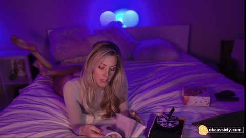 Media: Video of a blonde woman in a light sweater, lying on a bed, reading a book. Room is dimly lit with blue LED lights.